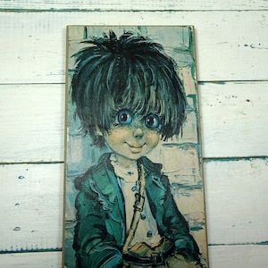 May include: A vintage print of a boy with blue eyes and dark hair wearing a green jacket and a white shirt. The print is on a wooden panel.