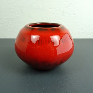 Op de afbeelding: Een rode keramische vaas met een glanzende afwerking en zwarte accenten. De vaas heeft een afgeronde vorm en een brede opening.