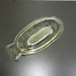 May include: A clear glass dish with a textured bottom and a handle. The dish is oval shaped and has a raised edge.