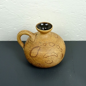 Può includere: Un vaso in ceramica marrone con un bordo marrone scuro e un design maculato. Il vaso ha un corpo arrotondato e un manico.