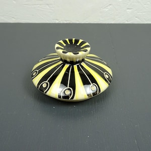 Può includere: Un coperchio in ceramica con un motivo a strisce nere e gialle. Il coperchio ha una piccola maniglia arrotondata con un motivo abbinato. Il design incorpora piccole forme ovali bianche con dettagli neri. Probabilmente per un vaso o contenitore decorativo.