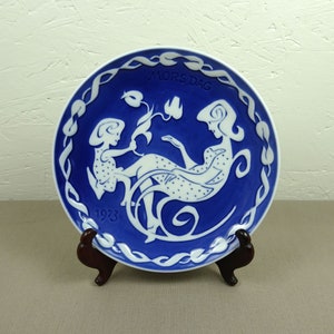 Pode incluir: Um prato de porcelana azul e branco com uma silhueta branca de uma mulher e uma criança. O prato tem uma borda branca com um desenho azul e as palavras "MORS DAG" e "1923" em branco.