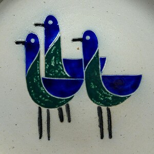 May include: A white ceramic plate with a stylized design of three blue and green birds. The birds are arranged in a circular pattern and have long, thin legs.