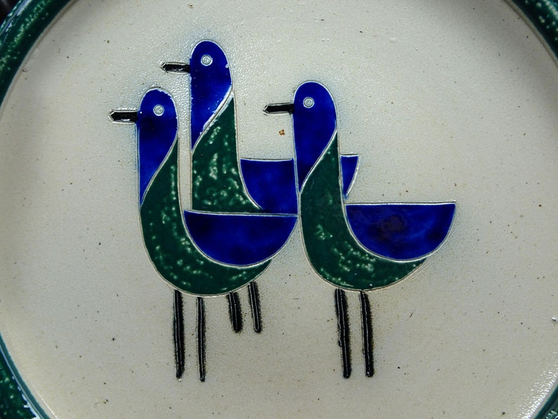 May include: A white ceramic plate with a stylized design of three blue and green birds. The birds are arranged in a circular pattern and have long, thin legs.