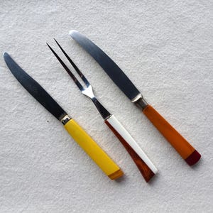 Può includere: Tre pezzi di posate con manici gialli, arancioni e bianchi. Le posate includono una forchetta, un coltello da burro e un coltello da tavola.