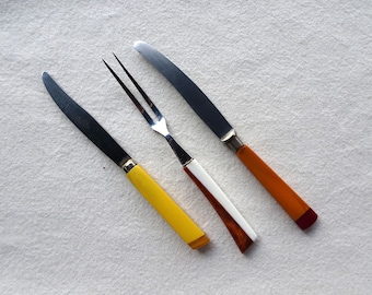 May include: Three pieces of cutlery with yellow, orange, and white handles. The cutlery includes a fork, a butter knife, and a table knife.