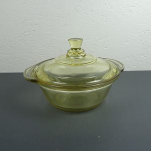 Op de afbeelding: Een vintage, lichtgele glazen ovenschaal met bijpassende deksel. De schaal heeft een afgeronde vorm met handvatten en een knop op het deksel. Het glas heeft een lichte groenachtige tint.