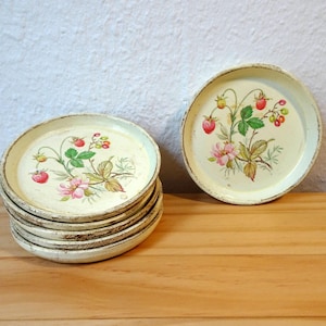 Könnte beinhalten: Ein Stapel von sechs cremefarbenen, runden Untersetzern mit einem verwitterten, gealterten Aussehen. Jeder Untersetzer zeigt ein Blumendesign mit Erdbeeren, rosa Blüten und grünen Blättern. Ein Untersetzer ist separat.