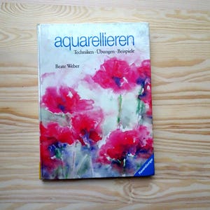 May include: A book titled "aquarellieren" with the subtitle "Techniken - Übungen - Beispiele" and the author's name, Beate Weber. The cover features a watercolor painting of red poppies. The book is on a wooden surface.