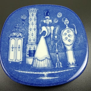 May include: A blue and white ceramic plate with a decorative design featuring a woman and a man in a room with a fireplace and other objects. The plate has a blue border with a white design.