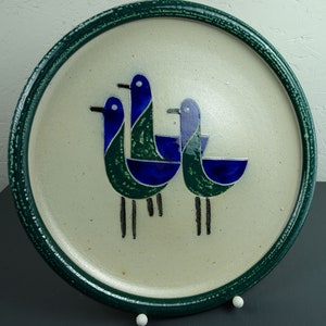 May include: A ceramic plate with a green rim and a white center featuring three stylized blue and green birds. The birds are arranged in a triangular pattern with their beaks pointing towards the center of the plate.
