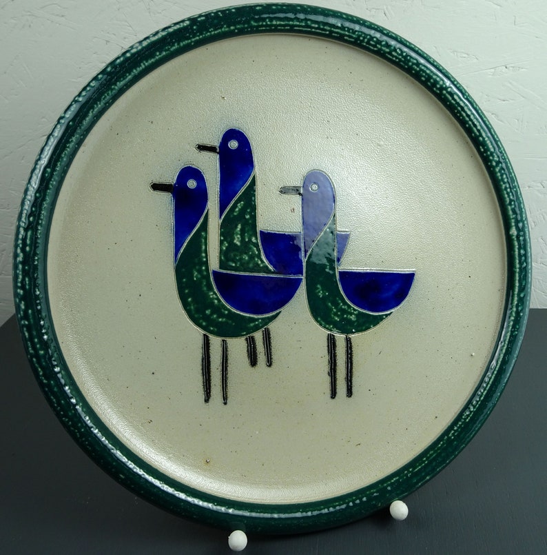May include: A ceramic plate with a green rim and a white center featuring three stylized blue and green birds. The birds are arranged in a triangular pattern with their beaks pointing towards the center of the plate.