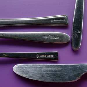 May include: Silverware set with various airline logos, including Swissair, Finnair, and Aero Lloyd, on a purple background. The set includes spoons and knives.