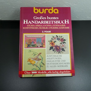 May include: A red book with the title "Großes buntes HANDARBEITSBUCH" and the text "2. FOLGE" on the cover. The book features illustrations of embroidery designs, including a floral wreath and a bird.