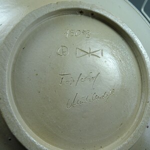 May include: Close-up of the bottom of a ceramic bowl with a white glaze. The bowl is marked with the number "150-B", a circle with a letter inside, a symbol that looks like a cross, the words "For/Schrof", and a signature that reads "Ulrich Wandbyl".