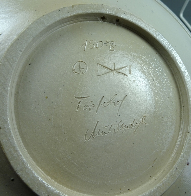 May include: Close-up of the bottom of a ceramic bowl with a white glaze. The bowl is marked with the number "150-B", a circle with a letter inside, a symbol that looks like a cross, the words "For/Schrof", and a signature that reads "Ulrich Wandbyl".