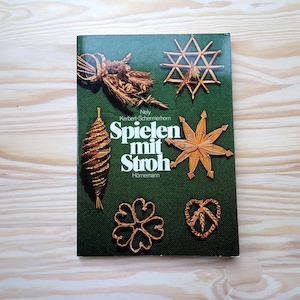 Könnte beinhalten: Buchcover mit dem Titel "Spielen mit Stroh" in weißer Schrift auf dunkelgrünem Hintergrund. Das Cover zeigt Strohhandwerk, darunter einen Stern, ein Herz und eine Tannenzapfenform. Das Buch liegt auf einer hellen Holzoberfläche.