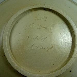 May include: The underside of a ceramic plate with a white glaze. The plate is marked with the number "450=B" and a signature that reads "Topfer" and "Ursula  Bauch".