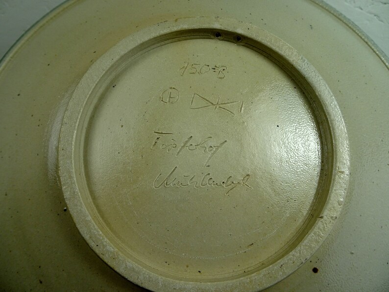 May include: The underside of a ceramic plate with a white glaze. The plate is marked with the number "450=B" and a signature that reads "Topfer" and "Ursula  Bauch".