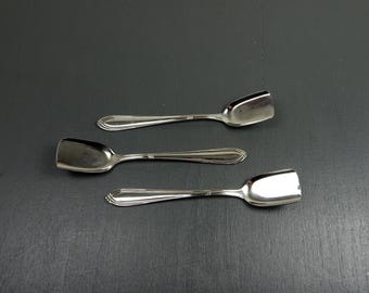 May include: Three silver serving spoons with rectangular bowls and decorative handles are arranged on a dark grey surface. The spoons are reflective, and the handles have a detailed design. The spoons are likely for serving sugar or other condiments.