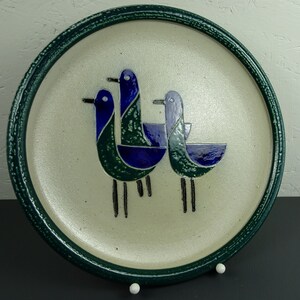 May include: A ceramic plate with a green rim and a white background features a stylized design of three blue and green birds.