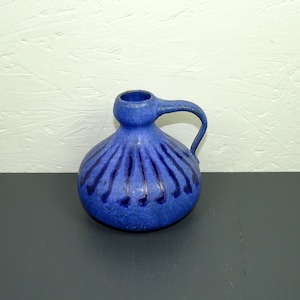 Può includere: Un vaso in ceramica blu con una superficie testurizzata e un manico. Il vaso ha un corpo arrotondato e un collo stretto.