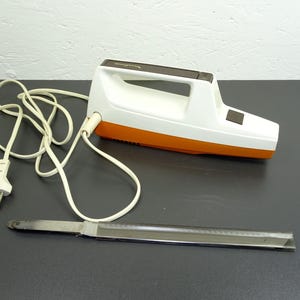 May include: An electric carving knife with a white and orange body, a brown handle, and a stainless steel blade. The knife is plugged into a wall outlet with a white cord. The knife is designed for slicing meats and other foods.