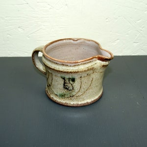 Può includere: Un piccolo brocca in ceramica fatta a mano con una glassa marrone e un manico marrone chiaro. La brocca ha una forma unica, leggermente asimmetrica, e un piccolo beccuccio.