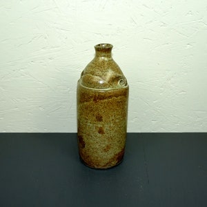 Może przedstawiać: Brązowa i beżowa ceramiczna butelka o zaokrąglonym korpusie i wąskiej szyjce. Butelka ma fakturowaną powierzchnię i mały uchwyt z boku.