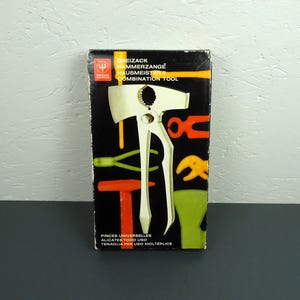 Op de afbeelding: Vintage combinatiegereedschap in de originele verpakking. De doos heeft een zwarte achtergrond met kleurrijke illustraties van gereedschap en de tekst "DREIZACK HAMMERZANGE HAUSMEISTER II COMBINATION TOOL". Het gereedschap is wit.