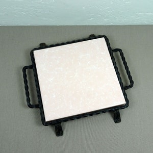 May include: A square ceramic tile with a white and pink marbled design is set in a black wrought iron frame with twisted rope detailing. The frame has four legs and two handles.