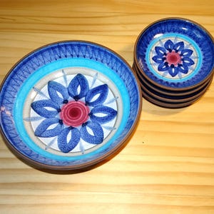 Op de afbeelding: Een set blauwe keramische borden met een bloemmotief. De borden hebben een centraal bloemontwerp in blauw, roze en wit, omringd door concentrische ringen van kleur. De borden zijn gestapeld en gerangschikt op een houten oppervlak.