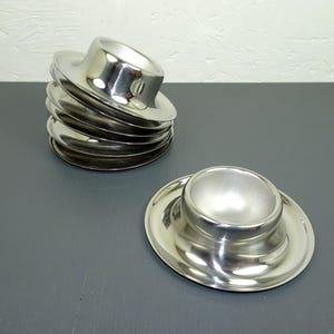 May include: A stack of six shiny, silver egg cups with a distinctive hat-like design, next to a single egg cup. The egg cups have a circular base and a rounded top, reflecting the light. The items are on a dark gray surface.