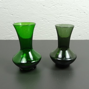 Könnte beinhalten: Zwei Glasvasen für Blumen, eine grün und eine dunkelgrün. Beide Vasen sind rund mit einem schmalen Hals.