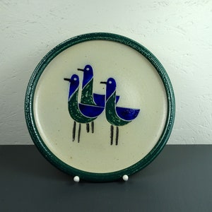 May include: A ceramic plate with a green rim and a white center featuring a stylized design of three blue and green birds.