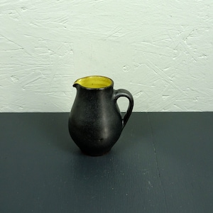 Può includere: Un piccolo brocca in ceramica nera con un bordo giallo e un manico. La brocca è su una superficie grigio scuro.