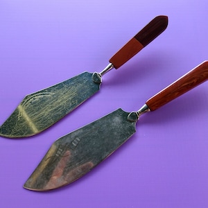 May include: Two silver cake servers with wooden handles. The handles are a dark brown color. The servers are on a purple background.