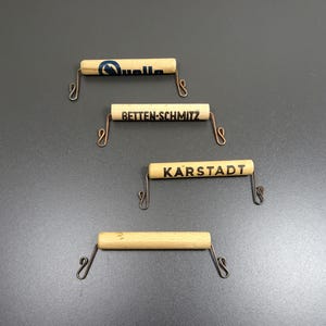 May include: Four wooden dowels with metal hooks on each end. The dowels are labeled with the names of different stores: "Quelle", "Betten-Schmitz", "Karstadt", and an unreadable name.