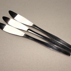 May include: Three silver butter knives laid out on a beige surface.