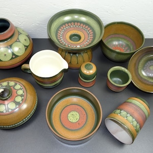 Può includere: Un set di 10 pezzi in ceramica in uno schema di colori verde, marrone e arancione con un motivo geometrico. Il set comprende un vaso, una brocca, una ciotola, una tazza, una lampada, un saliera e un piattino.