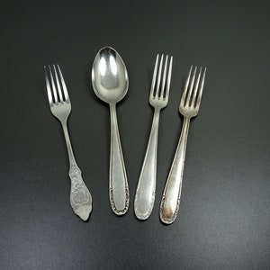 Può includere: Quattro posate, tra cui un cucchiaio e tre forchette, sono disposte su una superficie nera. Le posate sono in argento e hanno una finitura lucida.
