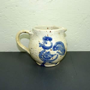 Può includere: Un piccolo brocca in ceramica bianca con un design di gallo blu. La brocca ha una glassa macchiettata e un corpo arrotondato.