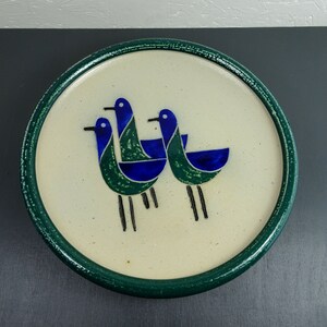 May include: A white ceramic plate with a green rim features a stylized design of three blue and green birds. The birds are depicted in a simple, geometric style.