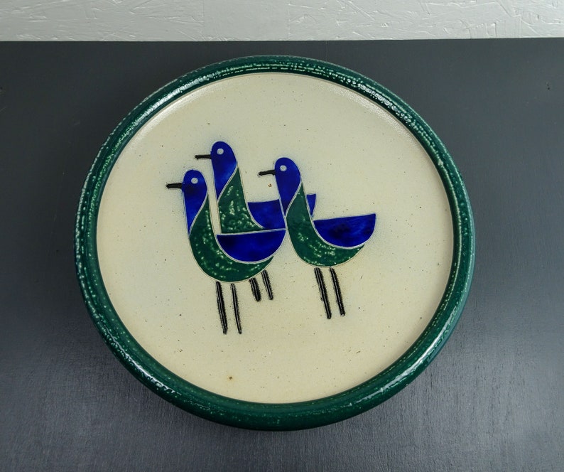 May include: A white ceramic plate with a green rim features a stylized design of three blue and green birds. The birds are depicted in a simple, geometric style.
