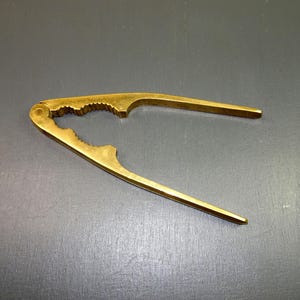 May include: A brass nutcracker with a lever design. The nutcracker is gold-coloured and has a textured gripping area. The handles are long and tapered, and the tool is open, ready for use. The background is a solid grey.