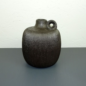 Può includere: Un vaso in ceramica marrone scuro con una superficie testurizzata e un piccolo manico. Il vaso ha una forma arrotondata e un'apertura leggermente svasata.