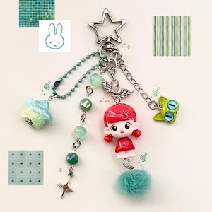 Puede incluir: Un llavero caprichoso con un cierre en forma de estrella y una variedad de dijes. Los dijes incluyen una muñeca pelirroja, una rana verde, una estrella y un pompón de color verde azulado. El llavero también tiene una cadena y detalles de cuentas en tonos verdes y plateados.
