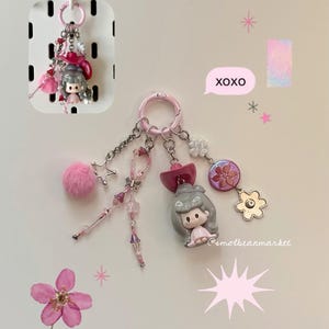 May include: A pink keychain with multiple charms, including a small doll, a fluffy pink pom-pom, and various beads and flowers. The keychain has a pink ring and several dangling chains. The text "XOXO" is in a speech bubble.