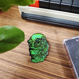 May include: Green enamel pin featuring a detailed illustration of the Creature from the Black Lagoon. The pin is on a wooden surface.