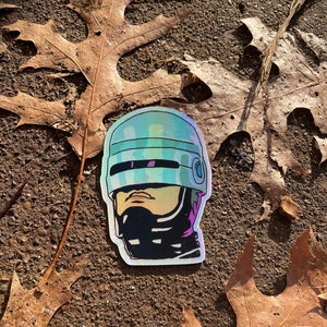 May include: A holographic sticker of a robot's head wearing a helmet. The helmet is blue and silver with a black visor. The robot's face is visible through the visor and has a serious expression.
